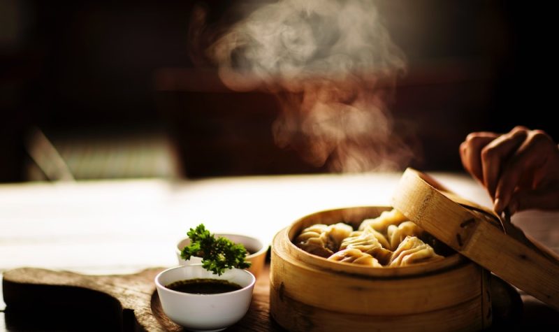 A hand is pulling the lid off a bamboo steam basket with dumplings inside while steam escapes.