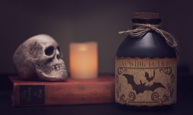 Un crâne d'humain et une bougie sont posés sur un livre épais à la couverture rouge. Une énorme fiole avec un bouchon en liège avec l'étiquette montrant un dessein de chauve souris et l'inscription potion d'aile de chauve-souris en anglais.
