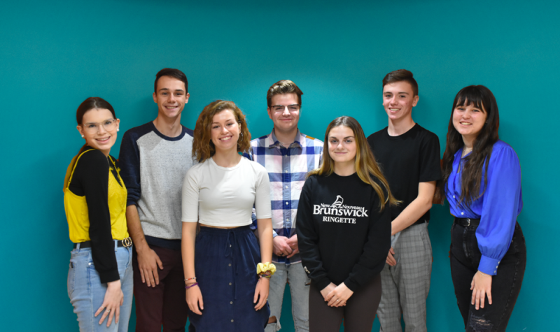 7 adolescents qui font partie de la Fédération des jeunes francophones du Nouveau-Brunswick (FJFNB) sont debout devant un arrière-plan bleu.