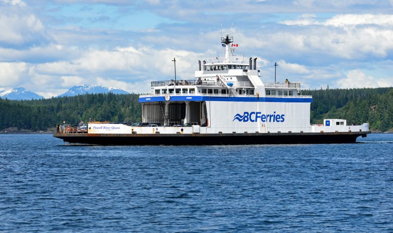 A white and blue ferry sails across a blue ocean under cloudy skies with forest in the background.