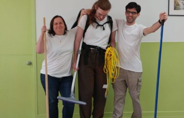 Three members of Powerhouse Co-Op wear matching white T-shirts and are holding cleaning equipment.