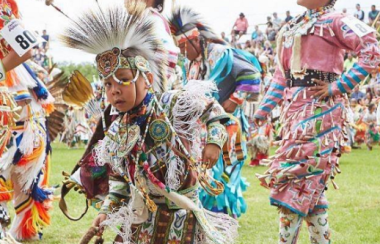 Child dancer in the foreground wearing traditional native american regalia dancing with female dancers also wearing the traditional native american regalia. Spectators look on in the background.