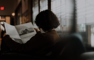 woman sitting on couch and reading the newspaper with her back facing us.