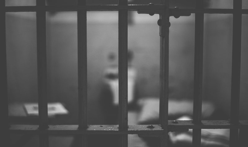 En noir et blanc, une grille de prison au premier plan. A l'arrière plan, une cellule floue on voir un lit.