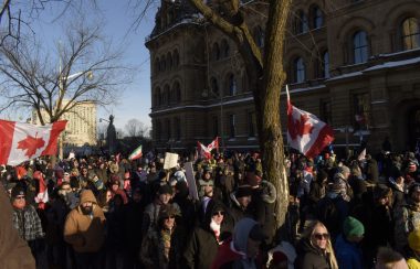 Many protestors are seen marching in downtown Ottawa, some with anti-vaccine signs and others with Canada flags, on a sunny winter day.