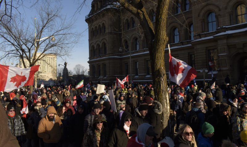 Many protestors are seen marching in downtown Ottawa, some with anti-vaccine signs and others with Canada flags, on a sunny winter day.