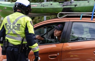 Photo of a police officer with a high-visibility jacket and a motorcycle helmet talking to a man in an orange car with a green kayak on the roof.