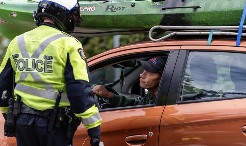 Photo of a police officer with a high-visibility jacket and a motorcycle helmet talking to a man in an orange car with a green kayak on the roof.