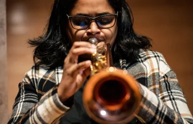 A dark haired woman wearing glasses and a black and brown plaid jacket facing the camera plays a trumpet.