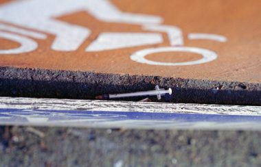On aperçoit une seringue pouvant avoir servi pour la consommation d'une drogue sur la bord d'une voie publique. On voit la chaussée et un symbole de circulation peint sur l'asphalte.