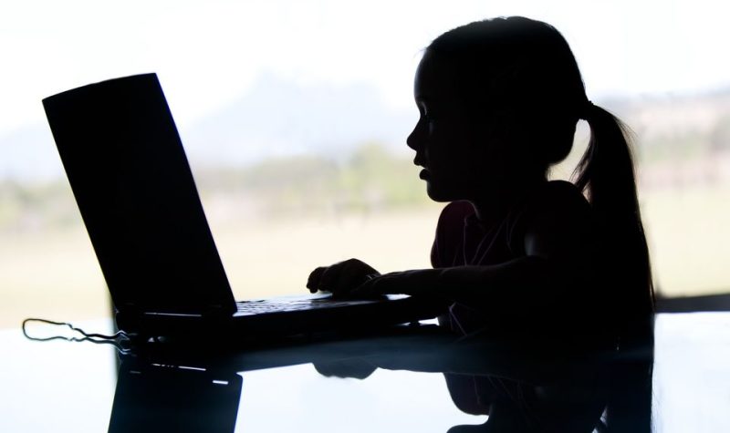 The dark silhouette of a young girl with a pony tail accessing a laptop.
