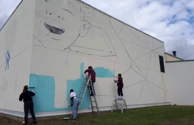 A photo of Facundo Gastiazoro and Stephanie Anderson painting a mural high on the outside wall of the recreational centre in Smithers.