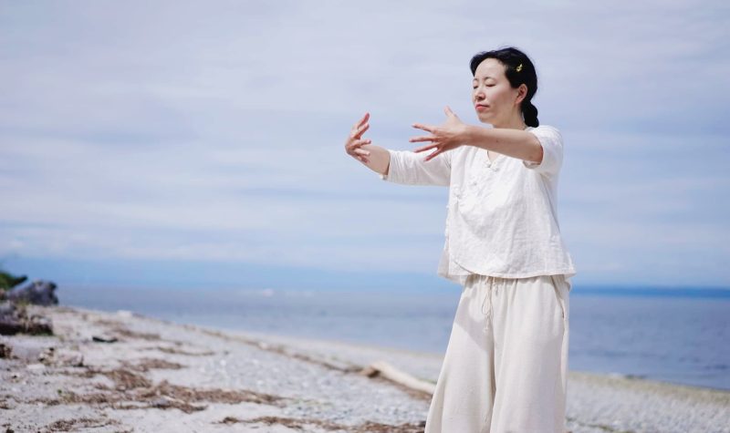 A woman holds a Tai Chi pose with her eyes closed on the beach.