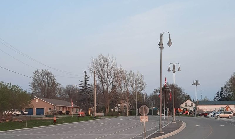 A road with a median and lightposts. Houses on the left. Cars in a parking lot for the ferry dock on the right. Trees in the background.