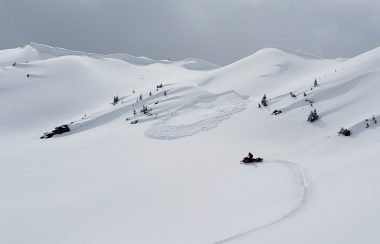 A wide shot of a ridge covered in snow, with a person on a snow mobile in the central left of the photo. A remote-controlled avalanche is seen being started, presumably by the person on the snow mobile. Weather is cloudy.