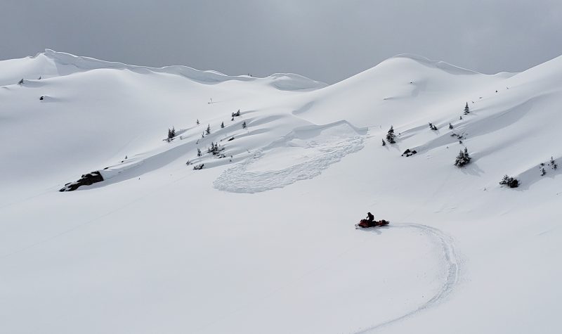 A wide shot of a ridge covered in snow, with a person on a snow mobile in the central left of the photo. A remote-controlled avalanche is seen being started, presumably by the person on the snow mobile. Weather is cloudy.