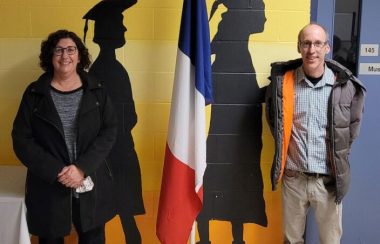 Une femme et un homme debout à chaque côté d,un drapeau acadien et en avant d'un mur jaune en briques avec dessins de finissants.