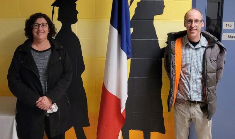 Une femme et un homme debout à chaque côté d,un drapeau acadien et en avant d'un mur jaune en briques avec dessins de finissants.