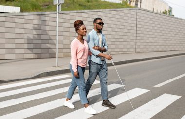 Two people are walking across a street together, one is holding a white cane indicating they are blind.