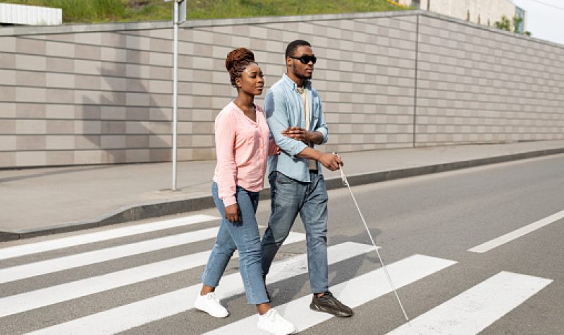 Two people are walking across a street together, one is holding a white cane indicating they are blind.