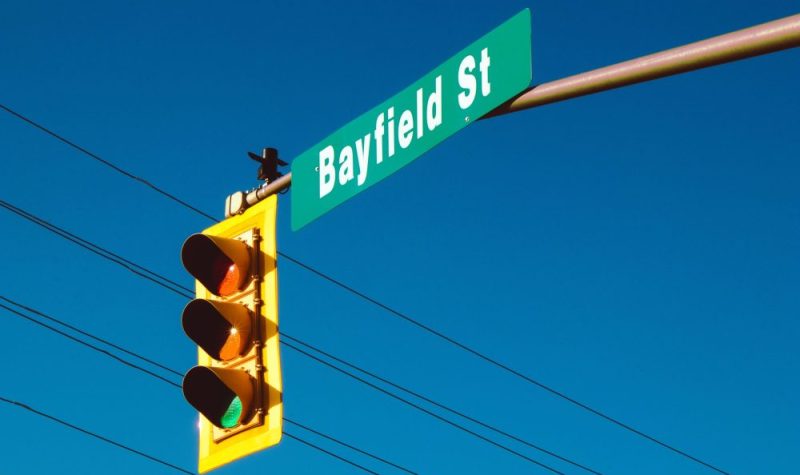 On aperçoit un feu de circulation avec un panneau de rue indiquant « Bayfield St ». À l'arrière plan on voit des fils électriques et le ciel bleu.