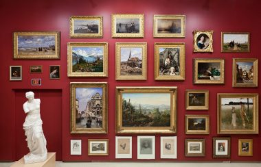 A red gallery wall covered in dozens of painting in gilded frames, and a white statue on a pedestal.