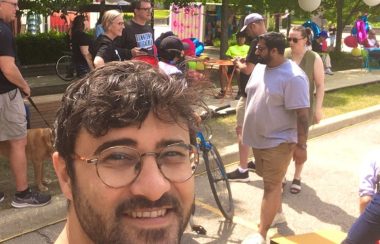 A bespectacled Sam Nabi takes a selfie at the Gaukel Block grand opening. People in the background are milling about. It is colourful and sunny.