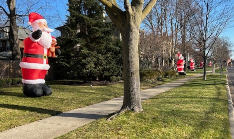 Inflatable Santa's can be seen standing in a row along a street in front of the sidewalk. Trees can also be seen throughout the neighborhood.