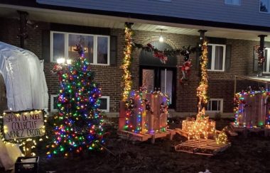 Vue d'ensemble d'un terrain décoré pour Noël. On trouve le sapin avec des lumières de Noël. À côté c'est écrit venez décorer. On voit aussi un rennes illuminées et deux petites cabanes à lutin avec des lumières de Noël.