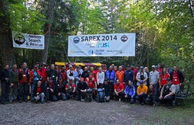 A group of people gathered in the woods. Trees and SAREX banners behind.