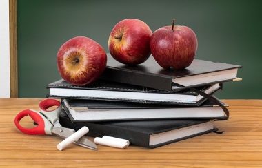 Trois pommes sont sur une pile de trois livres sur un bureau de bois du professeur avec une craie sur le côté. On voit un tableau vert en arrière-plan.