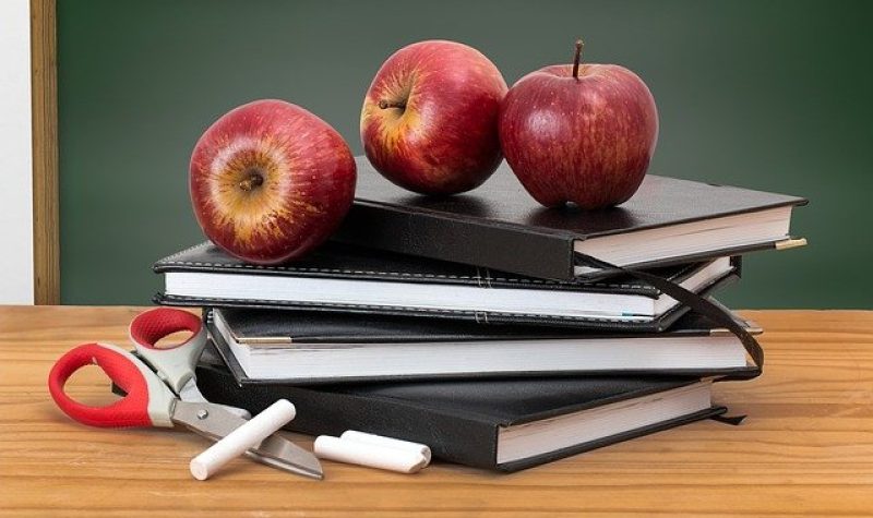 Trois pommes sont sur une pile de trois livres sur un bureau de bois du professeur avec une craie sur le côté. On voit un tableau vert en arrière-plan.