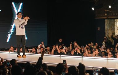 designer, Scott Wabano on a catwalk stage, being applauded by a crowd.
