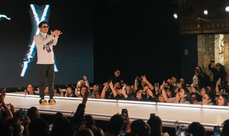 designer, Scott Wabano on a catwalk stage, being applauded by a crowd.