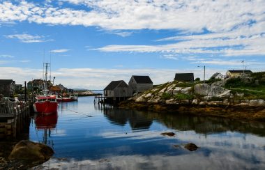 Un paysage sur l'eau avec un bateau et des habitations un peu au dessus du niveau de l'eau. Il y a du soleil et quelques nuages