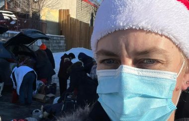 A woman wearing a blue face mask and a red Santa hat is seen from shoulders-up, looking at the camera. A vehicle with the trunk popped open is seen in the background, a group of people raking through it.