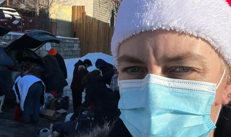 A woman wearing a blue face mask and a red Santa hat is seen from shoulders-up, looking at the camera. A vehicle with the trunk popped open is seen in the background, a group of people raking through it.