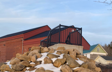 A platform and large pile of rocks sits in front of a red brick building. There is snow on the ground and a blue sky in the background.