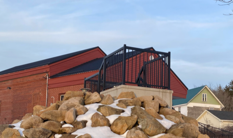 A platform and large pile of rocks sits in front of a red brick building. There is snow on the ground and a blue sky in the background.