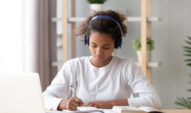 Une femme afro-américaine concentrée portant des écouteurs blancs, des notes d'écriture, des notes d'études avec un ordinateur portable et des livres. Il y a une étagère avec plantes à l'arrière.