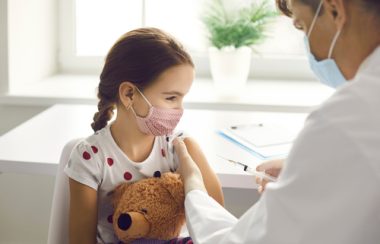 Jeune fille cheveux bruns coiffée d'une tresse, chandail blanc avec des pois rouges tenant un ours en peluche. Elle se fait vacciner par un médecin portant un sarrau blanc. Il y a une fenêtre derrière et une petite plante verte dans un pot blanc.