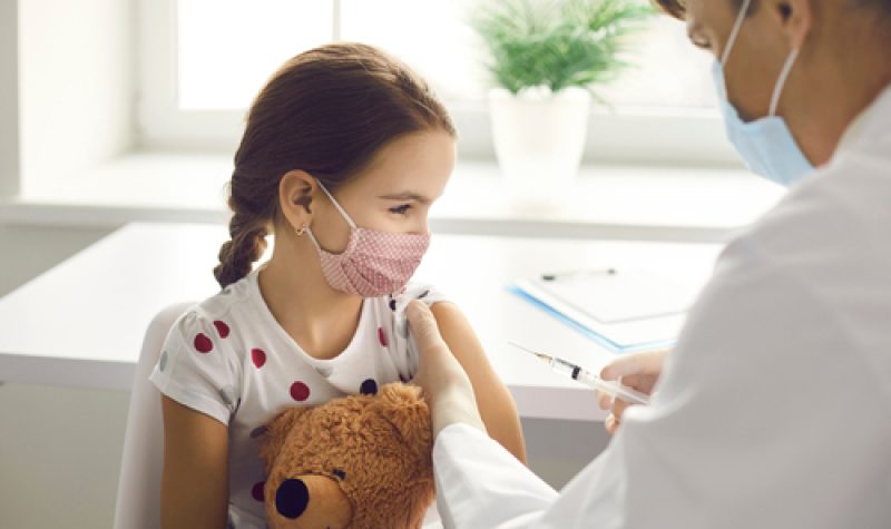 Jeune fille cheveux bruns coiffée d'une tresse, chandail blanc avec des pois rouges tenant un ours en peluche. Elle se fait vacciner par un médecin portant un sarrau blanc. Il y a une fenêtre derrière et une petite plante verte dans un pot blanc.