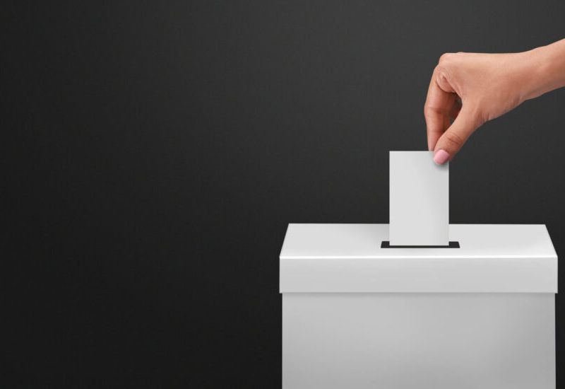 Femme déposant un papier de vote dans une boîte blanche sur fond gris foncé.