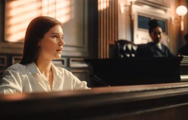 Femme à la cour de justice parlant devant un micro et observé par une juge. Le mobilier est brun foncé et les murs derrières blanc et beige.