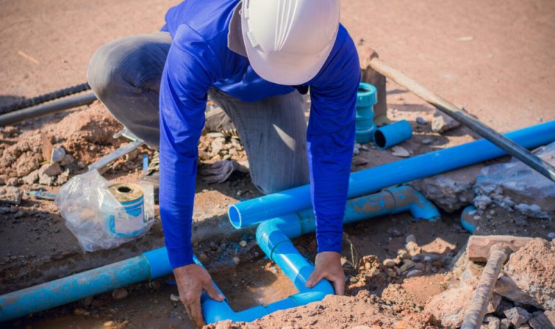 Homme portant un casque blanc, un chandail bleu et des pantalons gris travaille avec des tuyaux d'aqueduc bleus dans la terre.