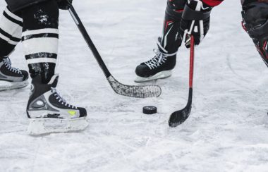 Sur une surface glacé, nous voyons les jambes avec équipement de hockeyeur avec une rondelle entre les deux.