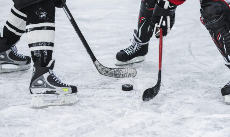 Sur une surface glacé, nous voyons les jambes avec équipement de hockeyeur avec une rondelle entre les deux.