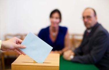 Femme avec chandail bleu et cheveux bruns à gauche et homme avec des lunettes, chauve et chemise grise. Ils sont assis et l'image est embrouillée. À l'avant une boîte de bois est sur une table avec une main qui dépose un papier blanc à l'intérieur.