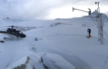 Justin Finn checks snow level beside a landed helicopter and a remote weather station in snowy mountains.