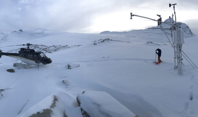 Justin Finn checks snow level beside a landed helicopter and a remote weather station in snowy mountains.